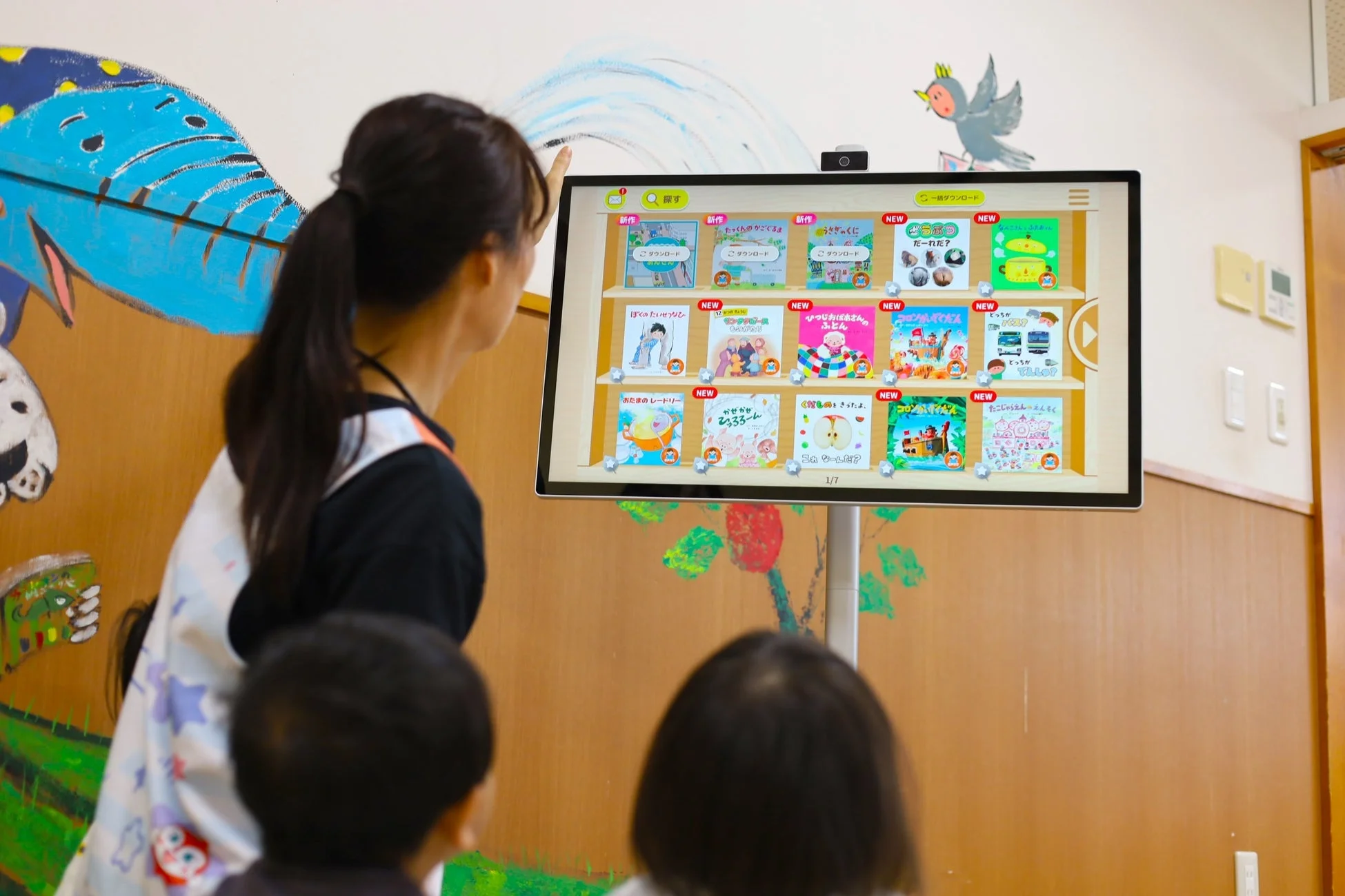 保育園か幼稚園で、先生が子どもたちに大型のデジタルディスプレイを使って絵本を選んでいる様子です。画面には多数の絵本の表紙が表示されており、インタラクティブな学習の場面が見られます。