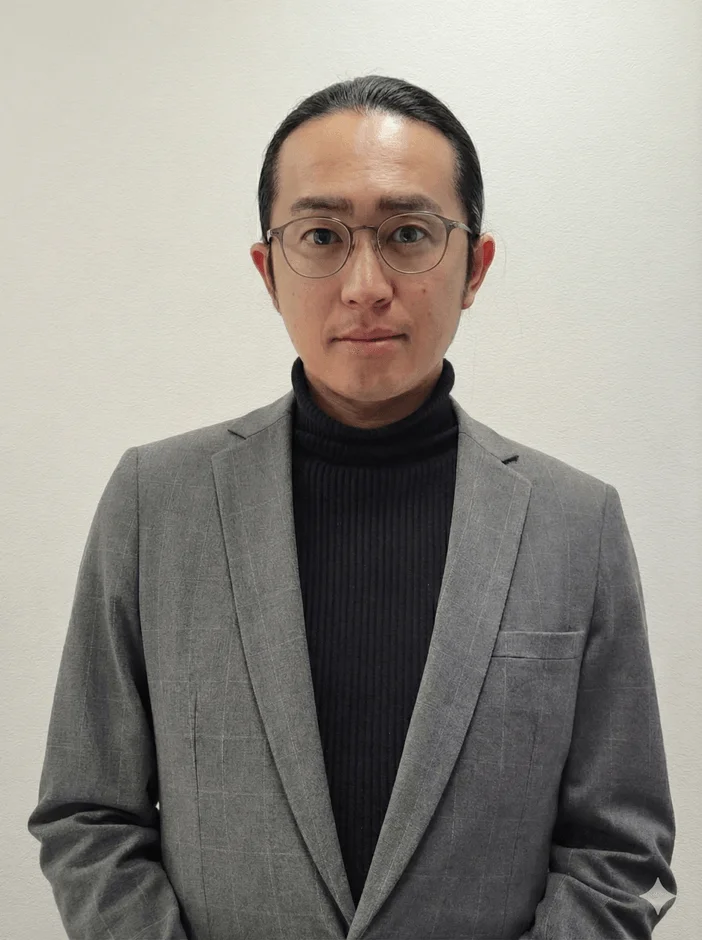 グレーのスーツジャケットと黒いタートルネックを着用し、眼鏡をかけた男性のポートレート。