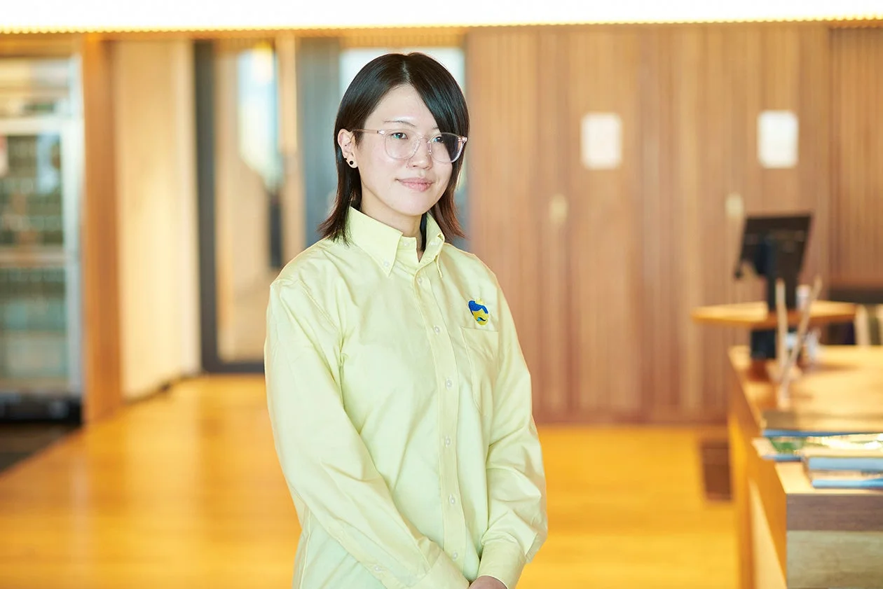 明るい室内に立つ若い女性のポートレート。眼鏡をかけ、黄色のシャツを着ており、胸元には青と黄色のバッジをつけている。穏やかな笑顔でカメラを見つめている様子。