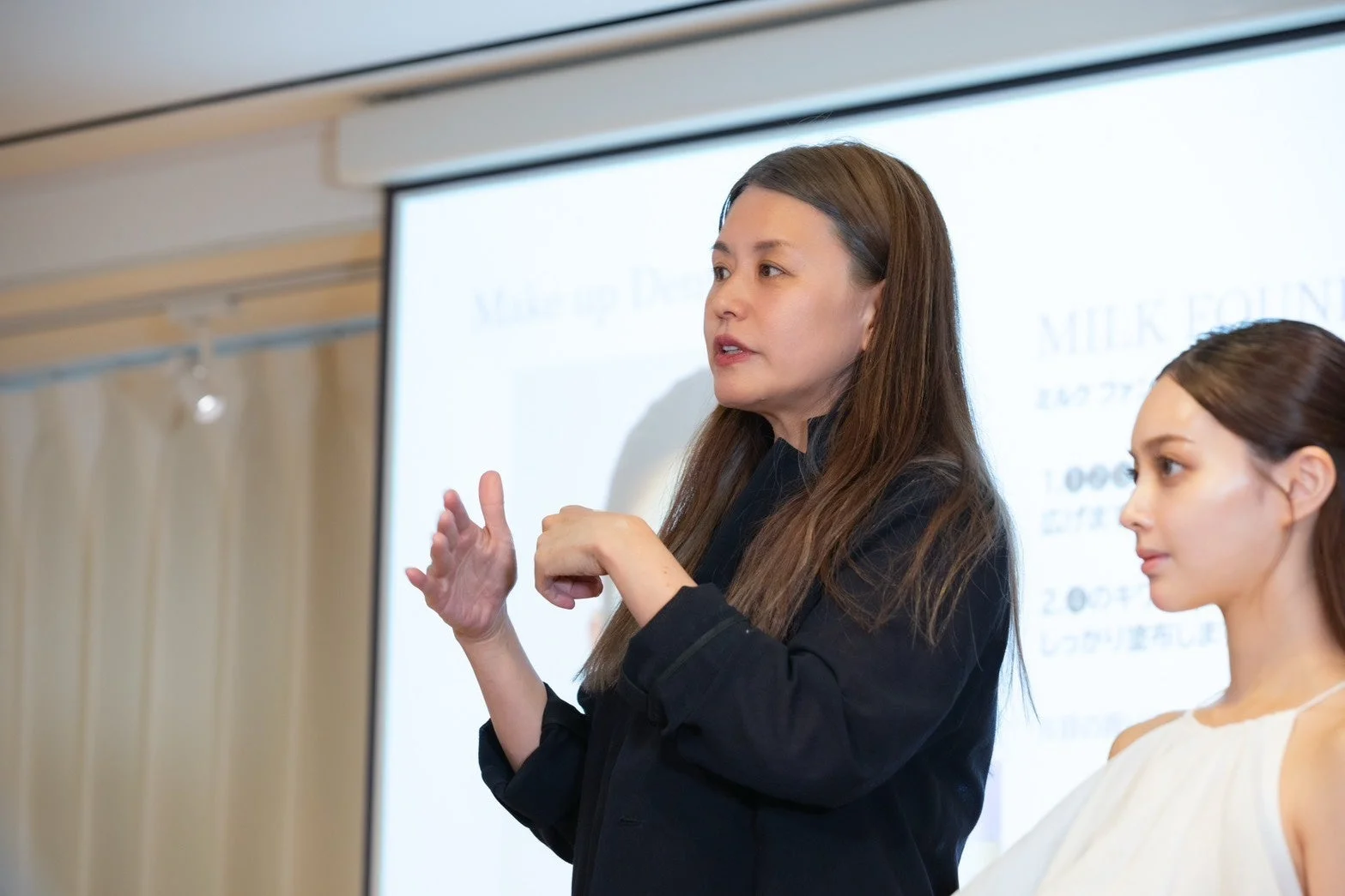 女性がステージ上でメイクアップに関するプレゼンテーションを行っており、スクリーンには「MILK FOUNDATION」の文字が見えます。